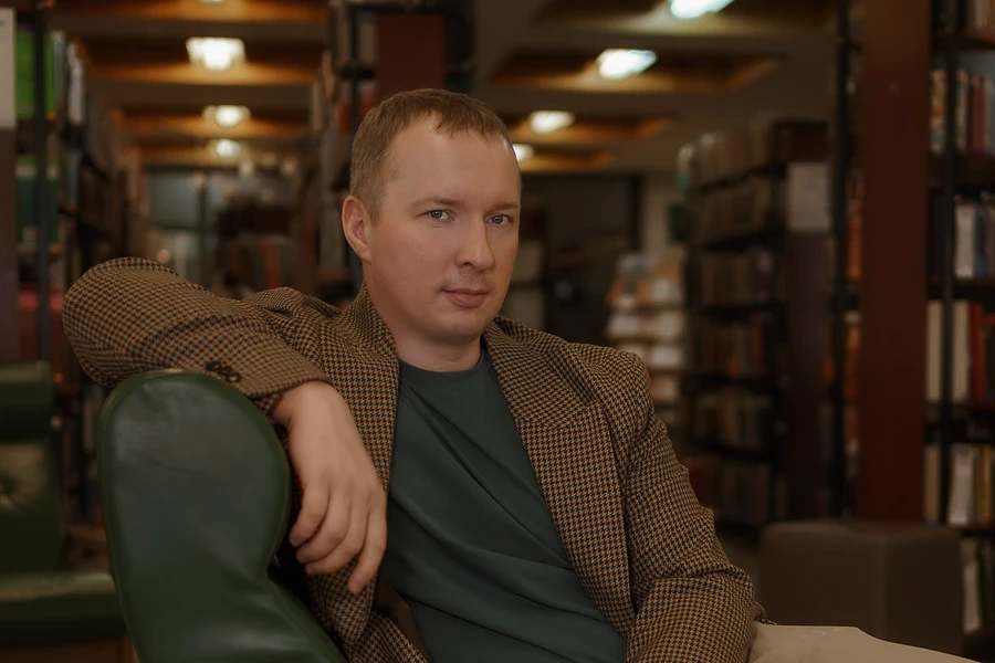 Денис Раченков в кресле на фоне книжных полок
